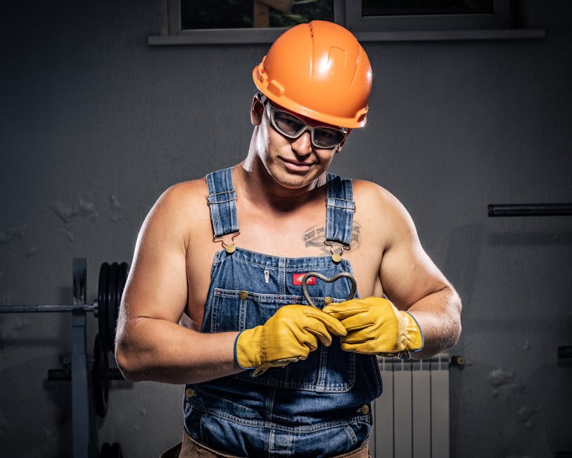 Muscular construction worker in overalls and helmet displaying strength and humor indoors.